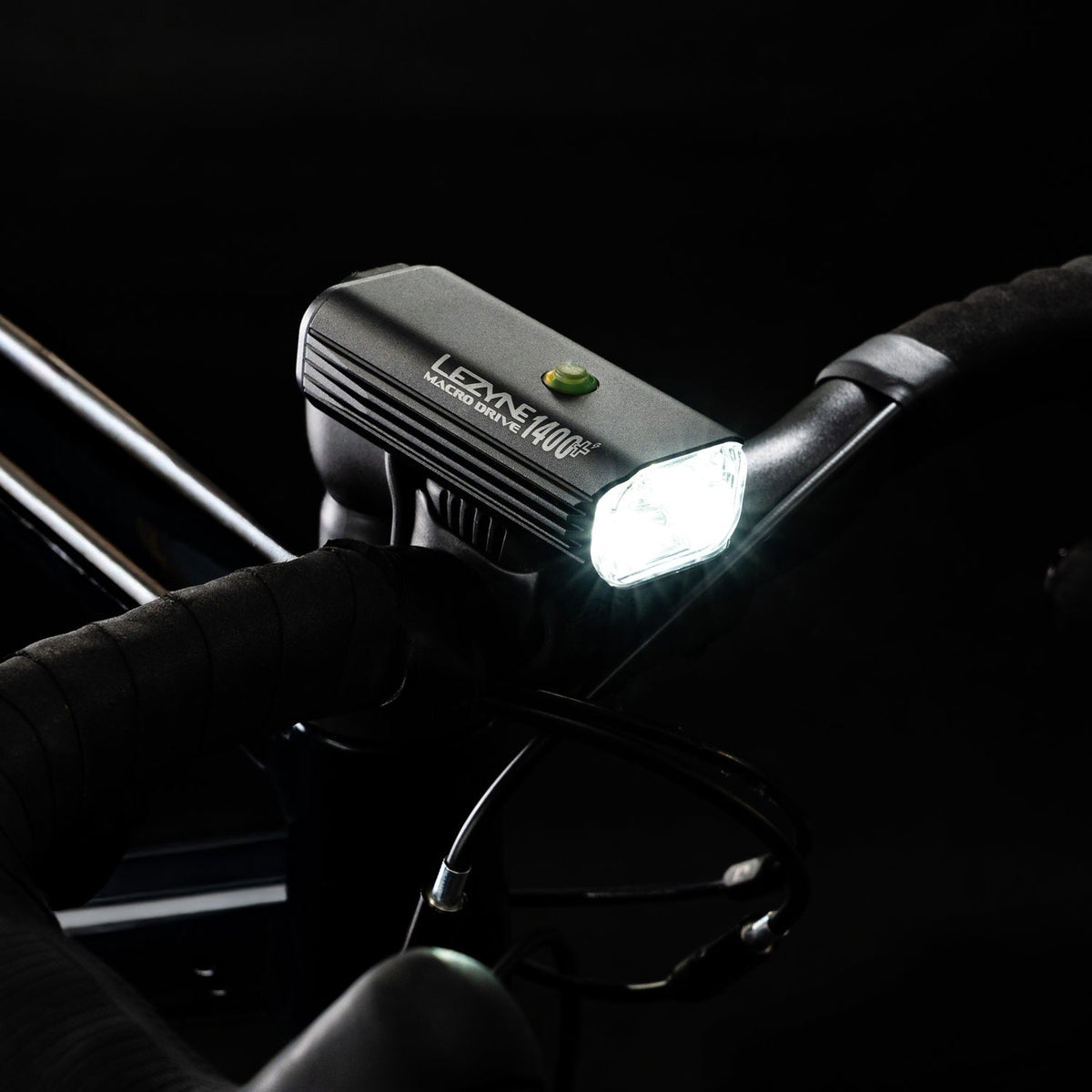 Lezyne Macro Drive 1400+ front light illuminated on bike handlebar, held by gloved hand in dark setting