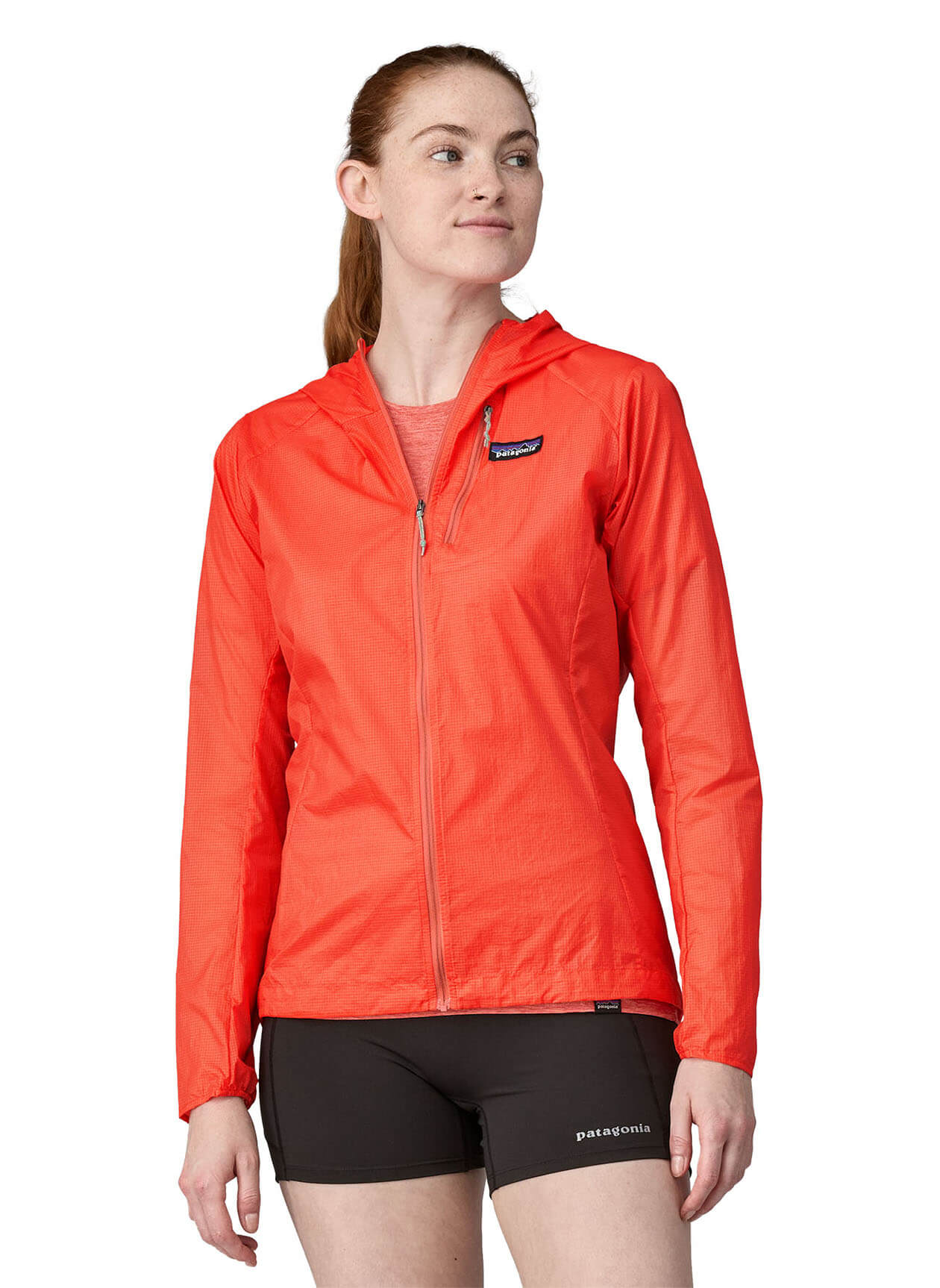 Woman wearing bright coral Patagonia Houdini jacket with black shorts on white background