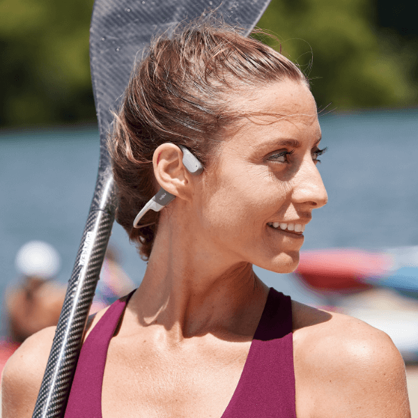 Woman wearing white AfterShokz bone conduction headphones while holding tennis racket outdoors