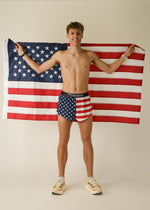 Man wearing ChicknLegs USA flag pattern split shorts holding American flag behind him