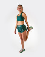 Woman wearing ChicknLegs forest green sports bra and matching shorts in stretching pose