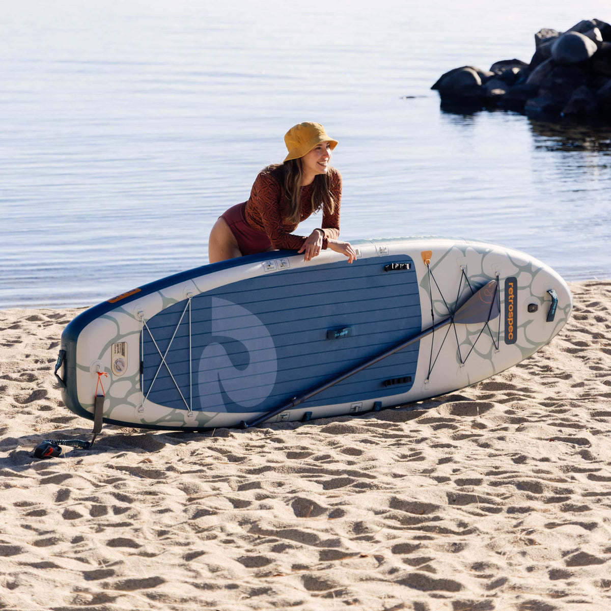 Woman in yellow hat leaning over Retrospec inflatable stand up paddle board on sandy beach