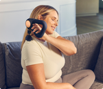 Woman using black Therabody Theragun Sense massage device on neck while sitting on couch