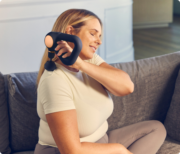 Woman using black Therabody Theragun Sense massage device on neck while sitting on couch
