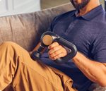 Man using black Therabody Theragun Sense percussion massager while sitting on couch