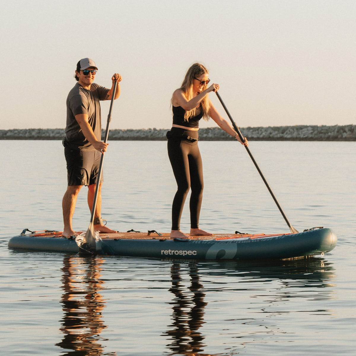 Two people stand-up paddleboarding on a Retrospec Weekender Crew inflatable SUP board at sunset