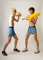 Two male models wearing ChicknLegs banana-print blue split running shorts playfully dueling with inflatable bananas