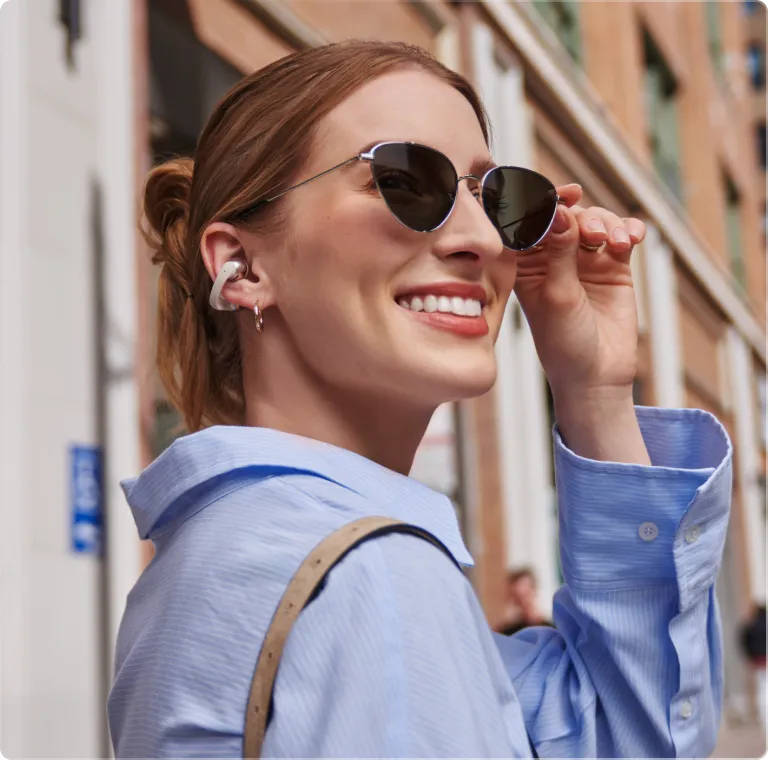 Woman wearing white Shokz OpenDots ONE wireless earbuds while adjusting sunglasses outdoors