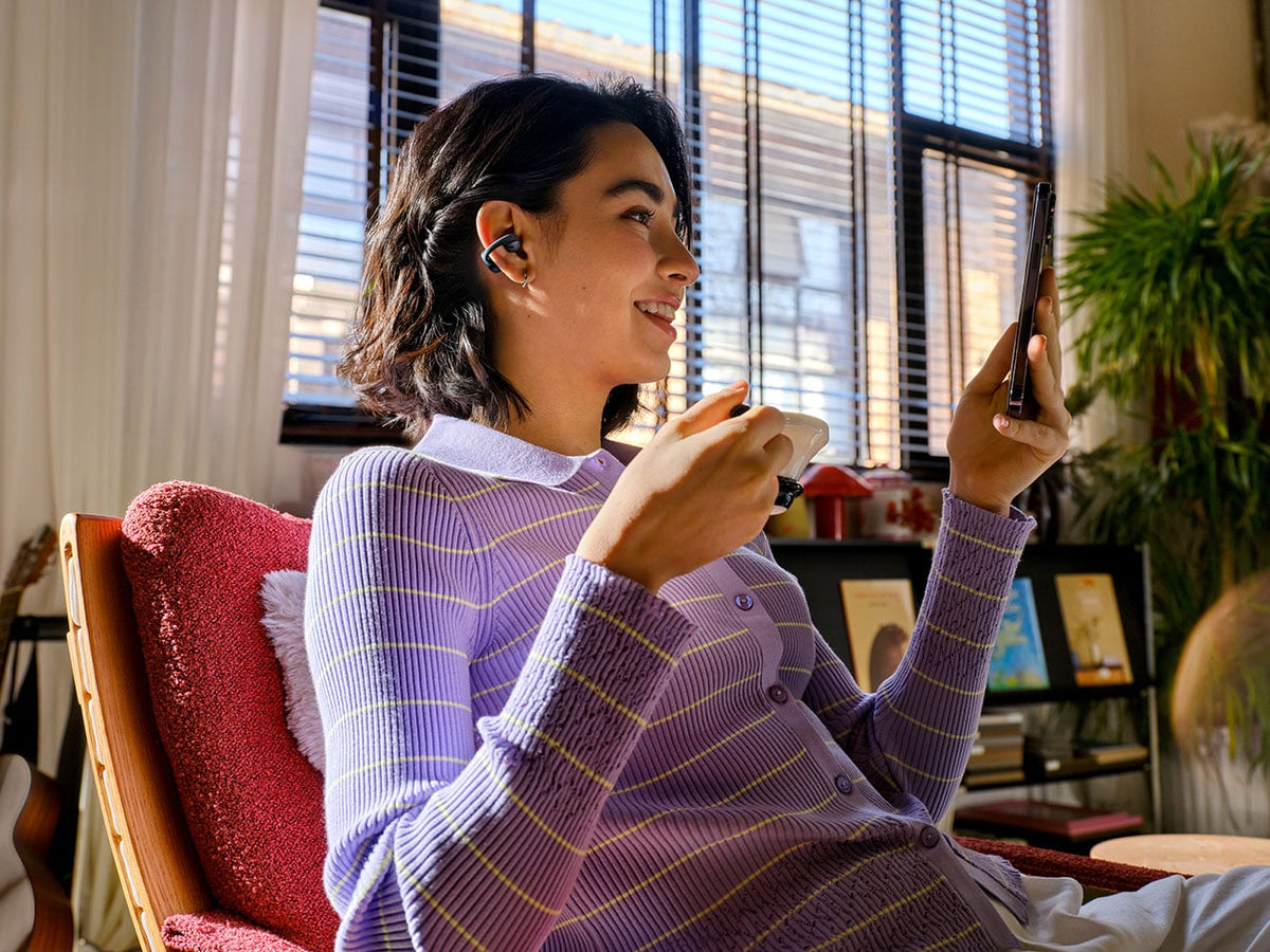 Woman wearing Shokz OpenDots Air black open-ear earbuds while looking at smartphone indoors